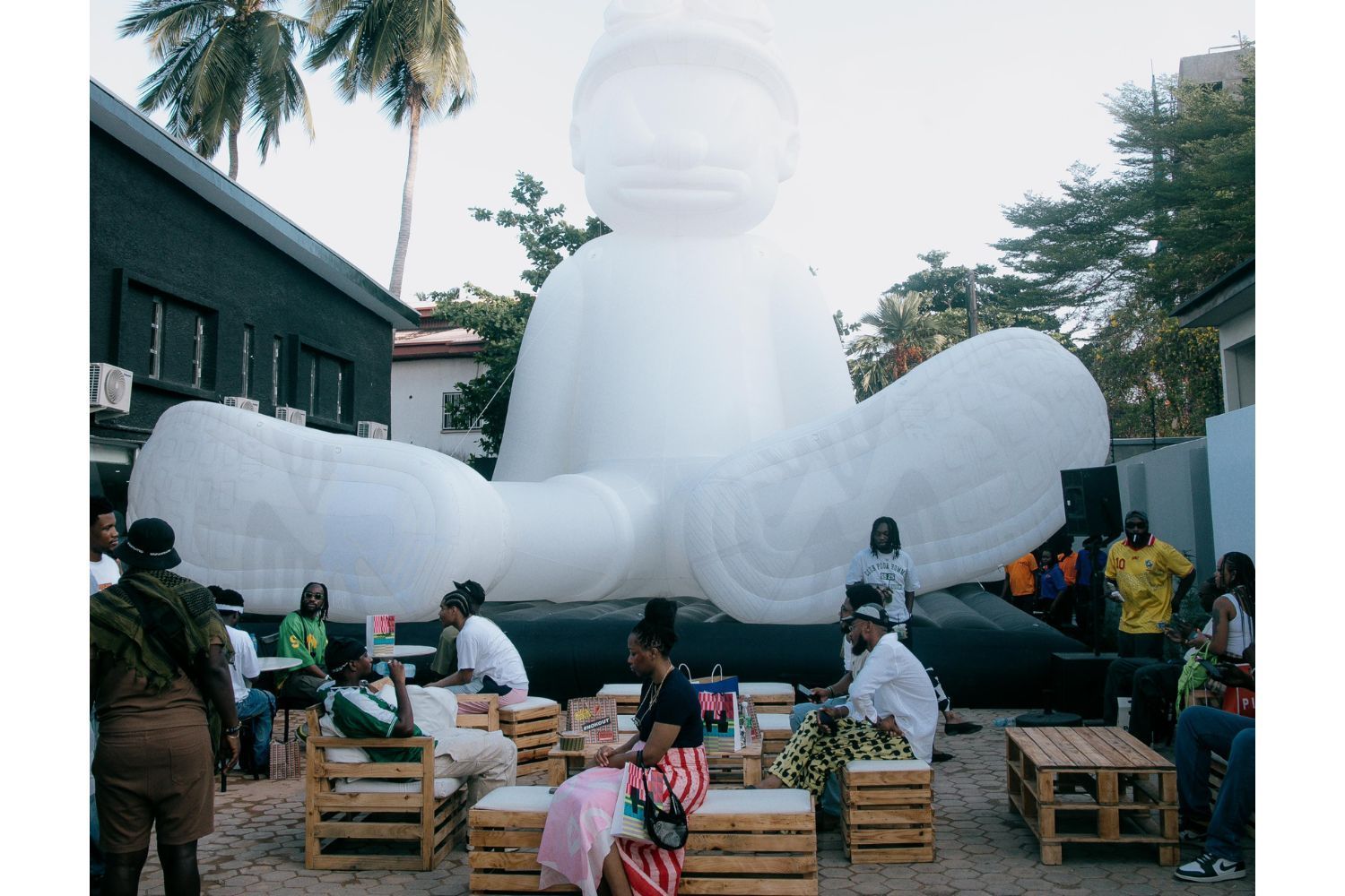 A group of people gather around wooden pallet seating in an outdoor courtyard, in front of a massive white inflatable figure with goggles, surrounded by palm trees and buildings.