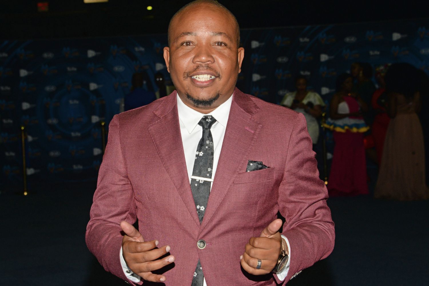 The late South African actor Presley Chweneyagae smiles at the camera. He is in a maroon suit and patterned tie poses for a photo at an indoor event, with people and a branded backdrop visible in the background.