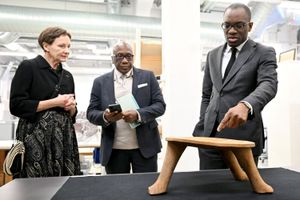 A man in a suit gestures toward a wooden three-legged object displayed on a table, while two other people, a woman and a man holding a phone and table, observe closely in a museum or archival setting.