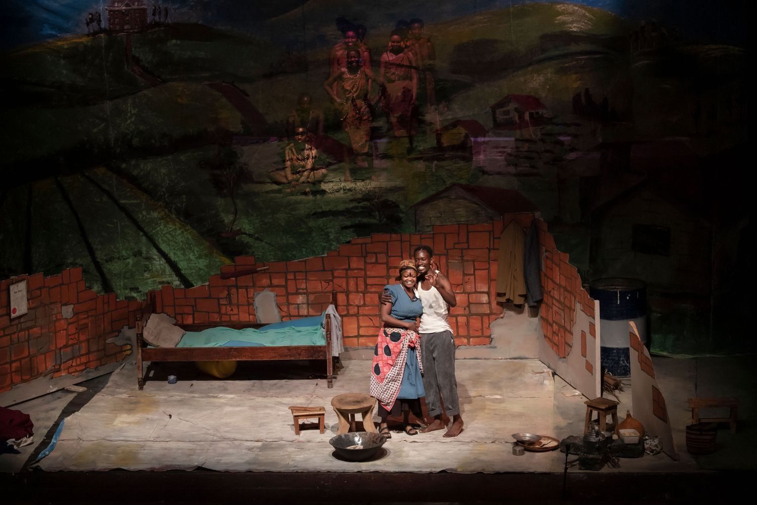 Two actors smile and embrace on a stage set that resembles a modest brick home, with painted backdrops of rural life and traditional African figures projected above them.