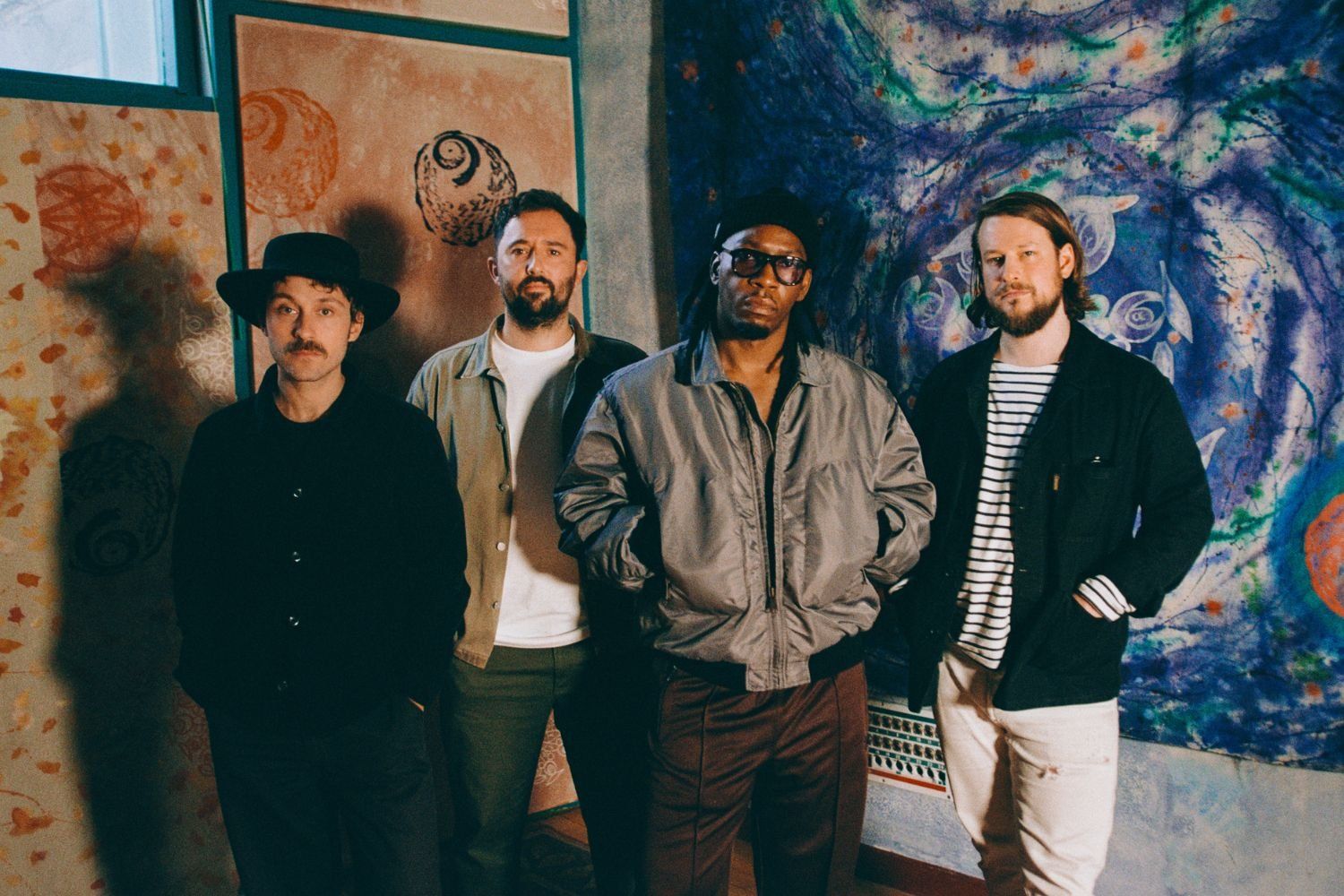 Four men,​ Tiggs Da Author and Thunderdrum, stand side by side in a warmly lit room, each with a distinct style—from casual jackets to a wide-brimmed hat—posed against a backdrop of colorful, abstract art panels.