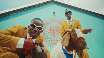 TitoM and Yuppe wearing yellow suits with silver white ties and holding on to two goats. 