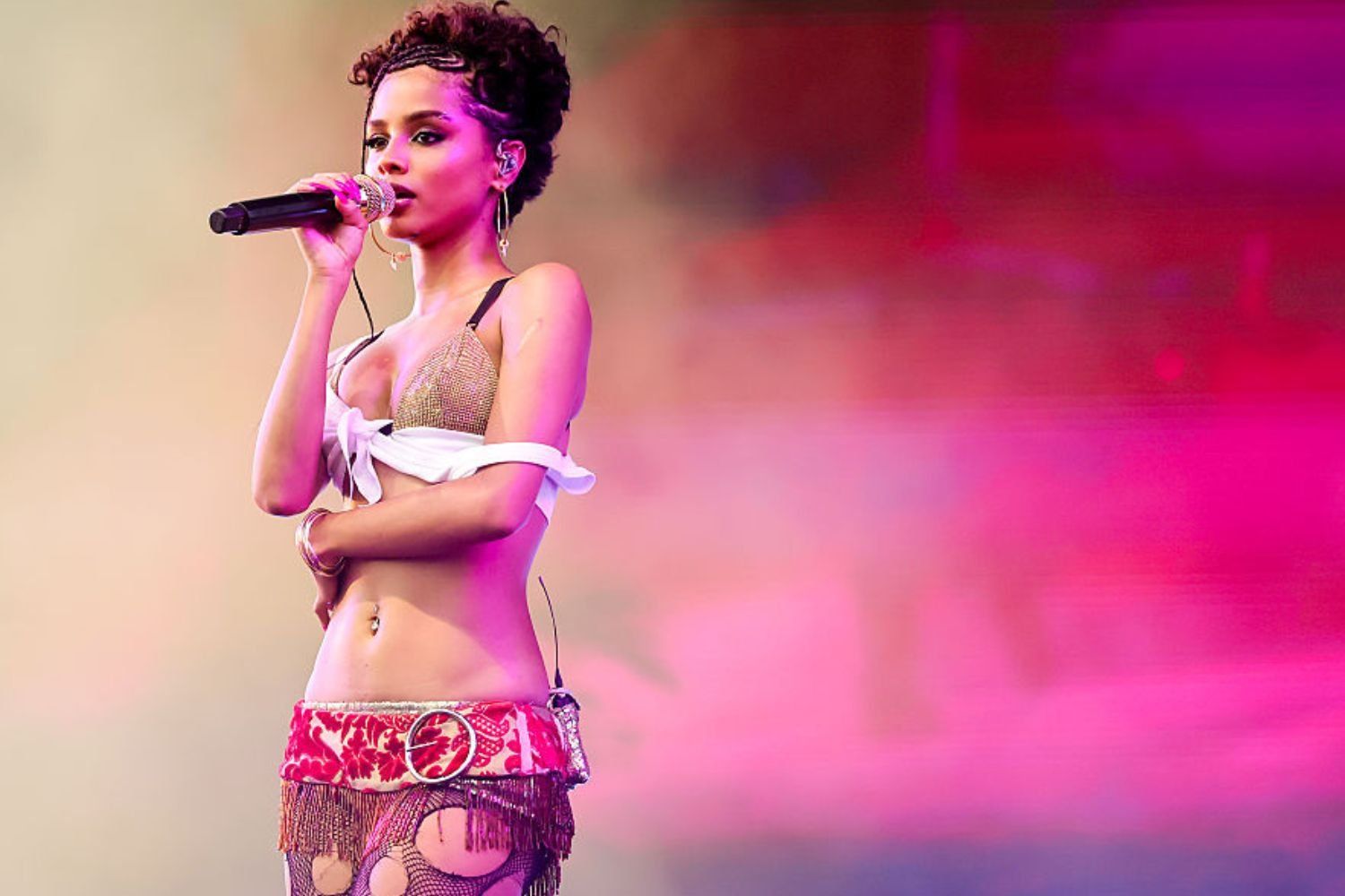 Tyla sings into a microphone on stage, wearing a shimmering bikini top, fishnet tights, and a red patterned hip scarf, set against a vibrant pink and purple background.