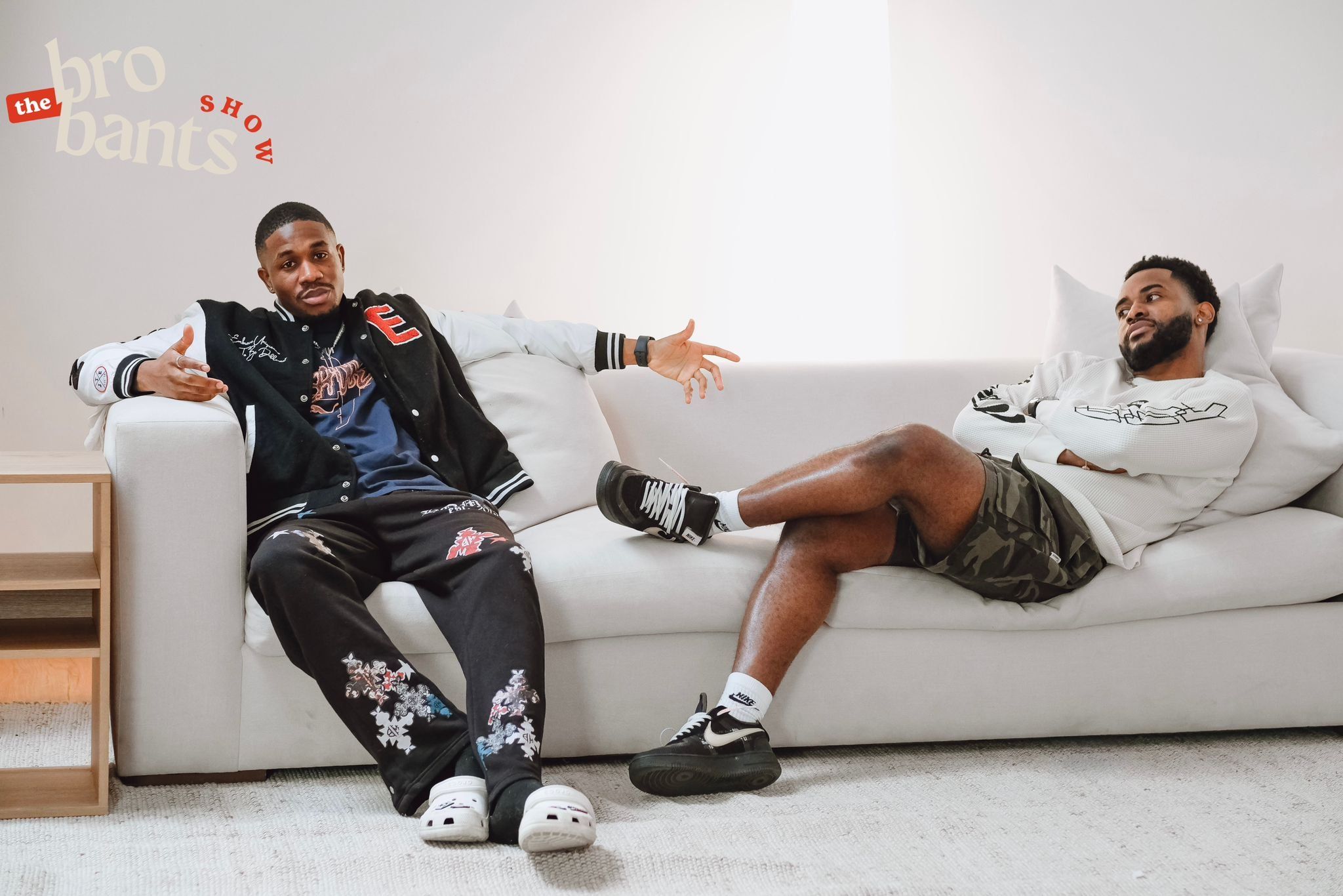 Two men are splayed out on opposite ends of a couch. One man, in a black and white varsity jacket and black jeans, is talking,  arms wide open, while looking at the camera. The other man, in shorts and a long-sleeved white top, is sitting with his arms and legs crossed, looking at the talking man. 