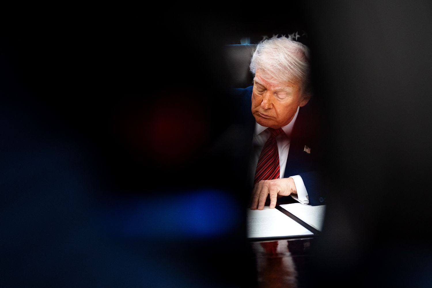 A dimly lit photo captures U.S. President Donald Trump signing a document at a desk, framed through a narrow, dark gap.