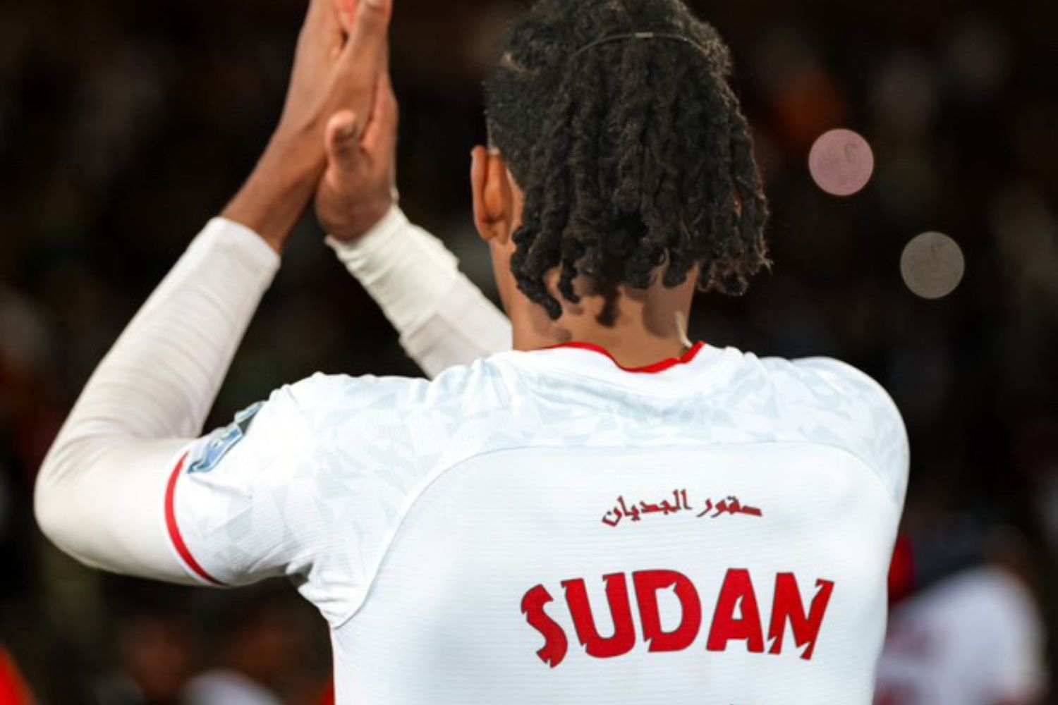 A football player is seen from behind, wearing a white jersey with red accents that reads “SUDAN” and the number 11. Arabic text is written above the country’s name. The player is clapping, under bright stadium lights, with blurred spectators in the background.