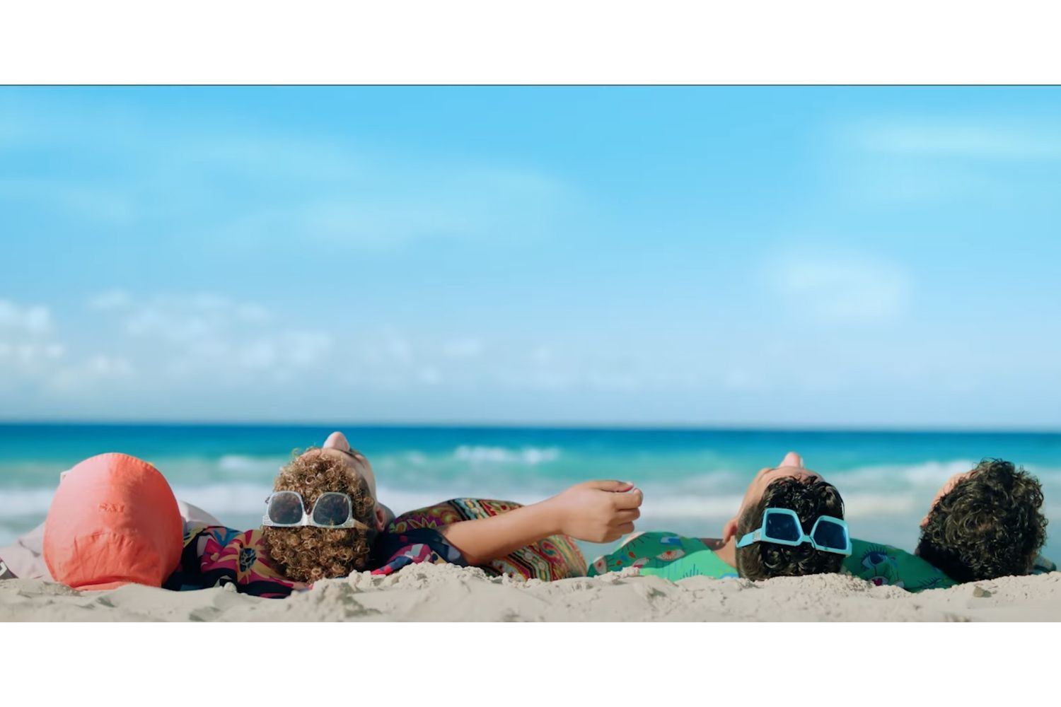 A group of four young men are lying on the beach, the turquoise  Mediterranean sea in the background and only the back of their heads are showing. Two of them have 