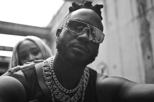 Black and white photo of Bebe Cool wearing chunky chains and sunglasses, with a woman in the background during a video shoot.