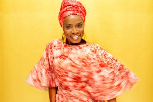 Angelique Kidjo poses in a red outfit and large gold earrings.  She wears a wide smile.