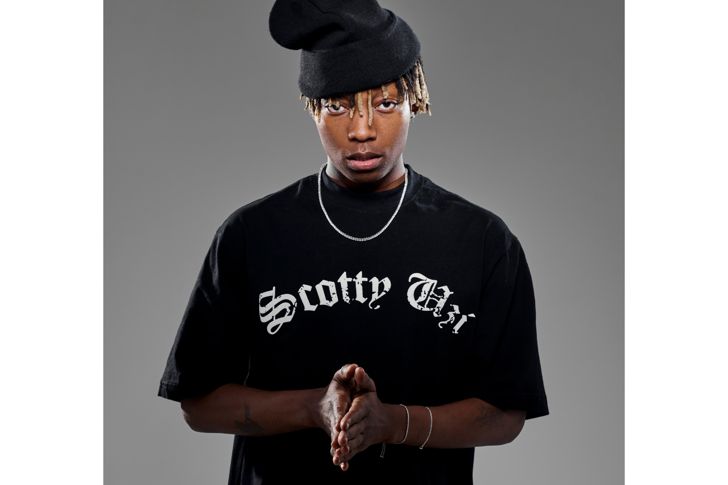 Xduppy stands against a plain gray background, gazing directly into the camera with a serious expression. He wears a black oversized T-shirt that reads “Scotty Uzi” in bold gothic script and a slouchy black beanie tilted to one side, partially covering his bleached-tipped locks. His hands are clasped together in front of him, and he accessorizes with a silver chain necklace and matching bracelets.