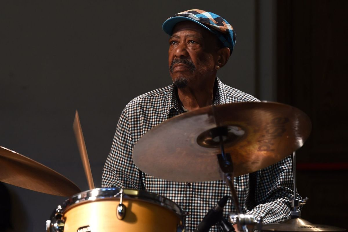 Louis Tebogo Moholo wears a checkered shirt and patterned cap and plays the drums, focused under soft stage lighting.