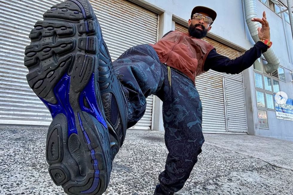Man Like B puts his best foot forward in a promo image. He wears black sneakers with a blue outline, navy blue pants with prints on them, and a stylish, light brown sleeveless jacket.