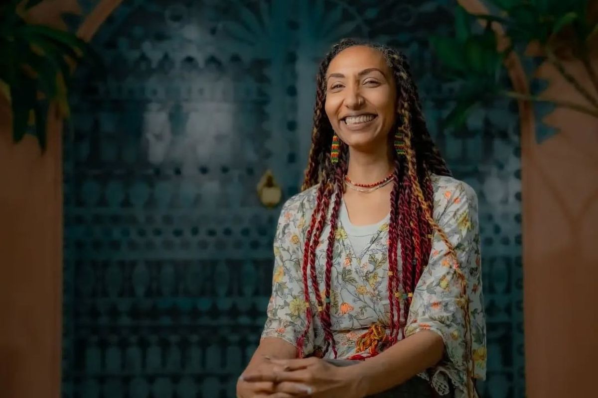 Maya Gadir sits legs crossed and hands on one knee, she is smiling brightly, hair braided in multicolored braids.