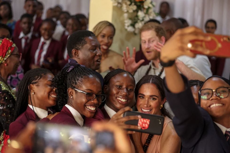 Meghan Markle takes a selfie with students with Prince Harry posing in the background.