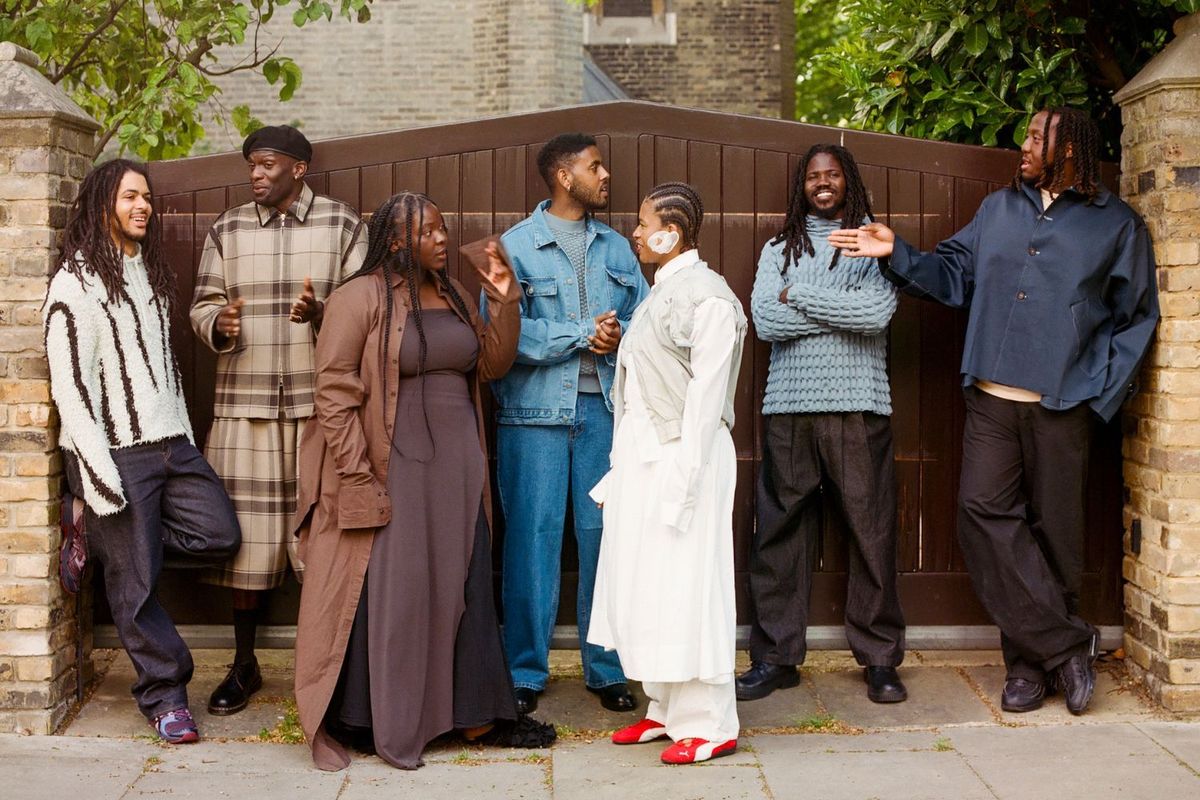 Members of the seven-piece outfit Kokoroko pose in front of a wooden entrance. They are in conversation, dressed stylishly in earthy tones.