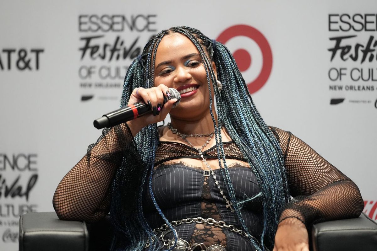 Moliy smiles and speaks into a mic; she is seated on a black leather chair, her hair in braids, in the background, the words: Essence Festival of Culture