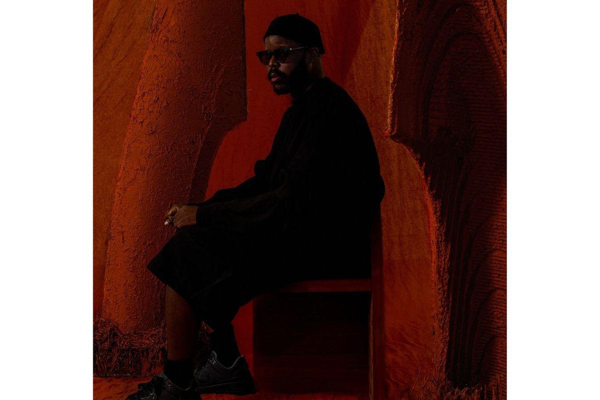 Myles Igwebuike photographed in dim light wearing a black outfit and sitting sideways.