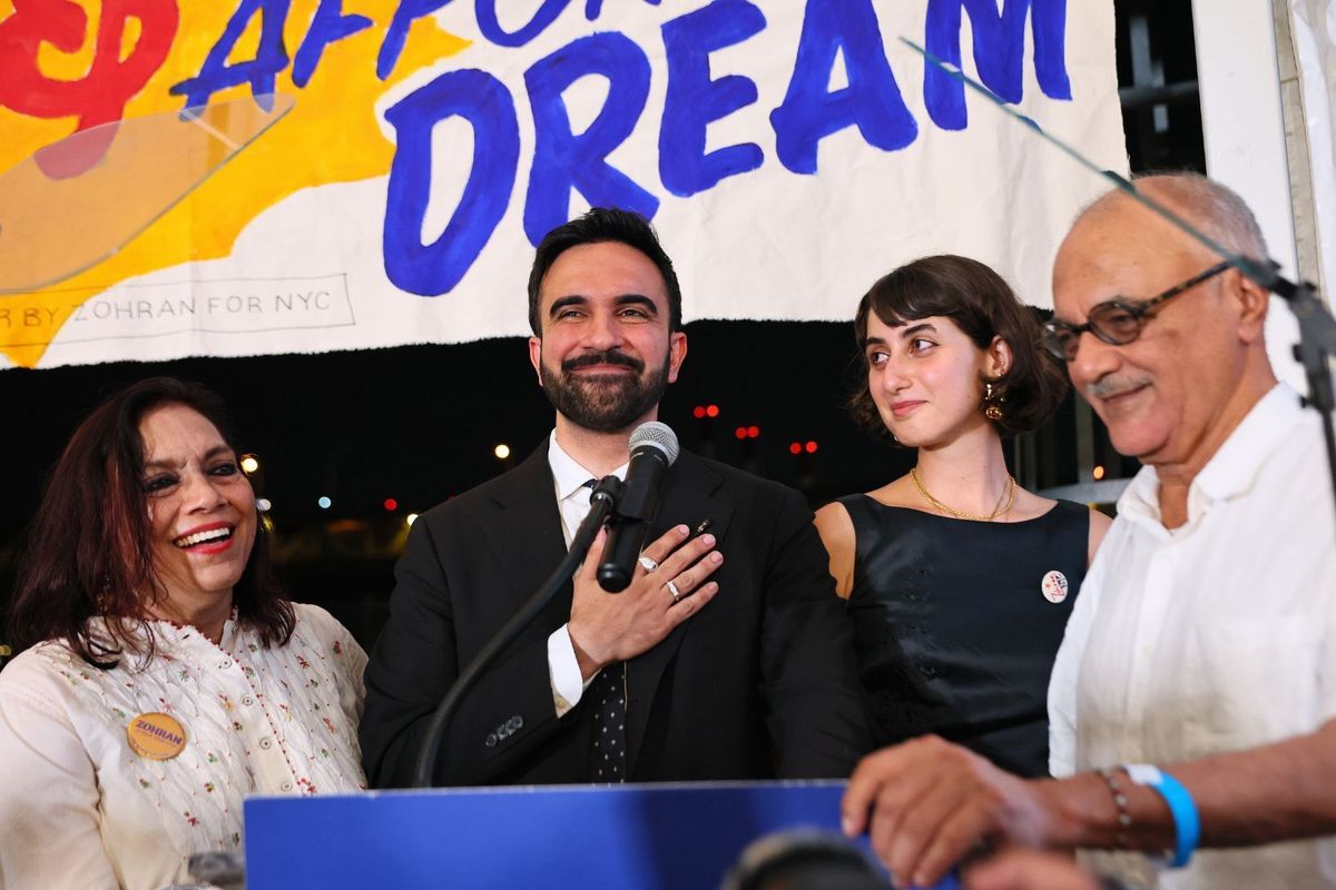 New York City mayoral candidate Zohran Mamdani stands in front of a mic smiling with a hand on his chest, his wife smiles ooks at him, his parents are in the picture also smiling.