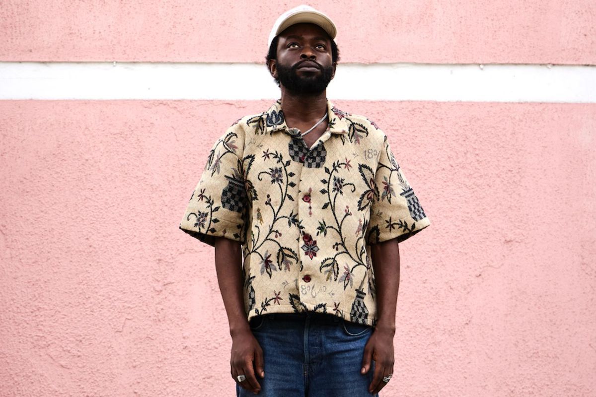 Nigerian artist Tim Lyre poses outdoors for a press shot. He’s in front of a peach pastel colored wall.