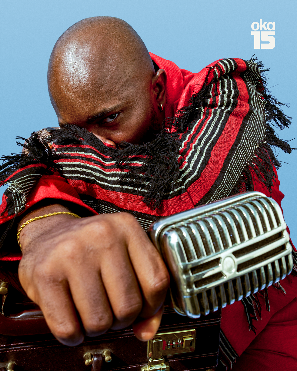 Nigerian music journalist is holding a microphone in the foreground of the frame, looking out with one eye, while his arm covers his other eye, and he's wearing a black and red suit with tassels.