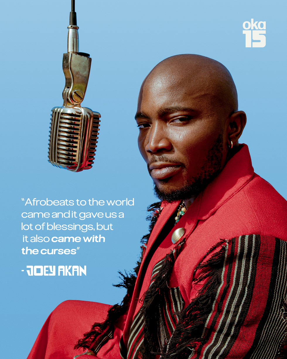 Nigerian music journalist Joey Akan is facing forward with his eyes looking towards the camera, with a microphone in front of his mouth, wearing a red and black suit with tassels on the front.