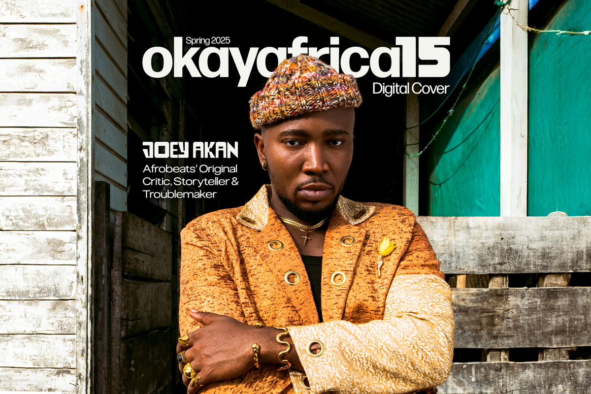Nigerian music journalist Joey Akan standing in front of a building with his arms crossed, wearing a yellow and gold jacket and a multicolored beanie-hat, and he has a determined, firm look on his face.