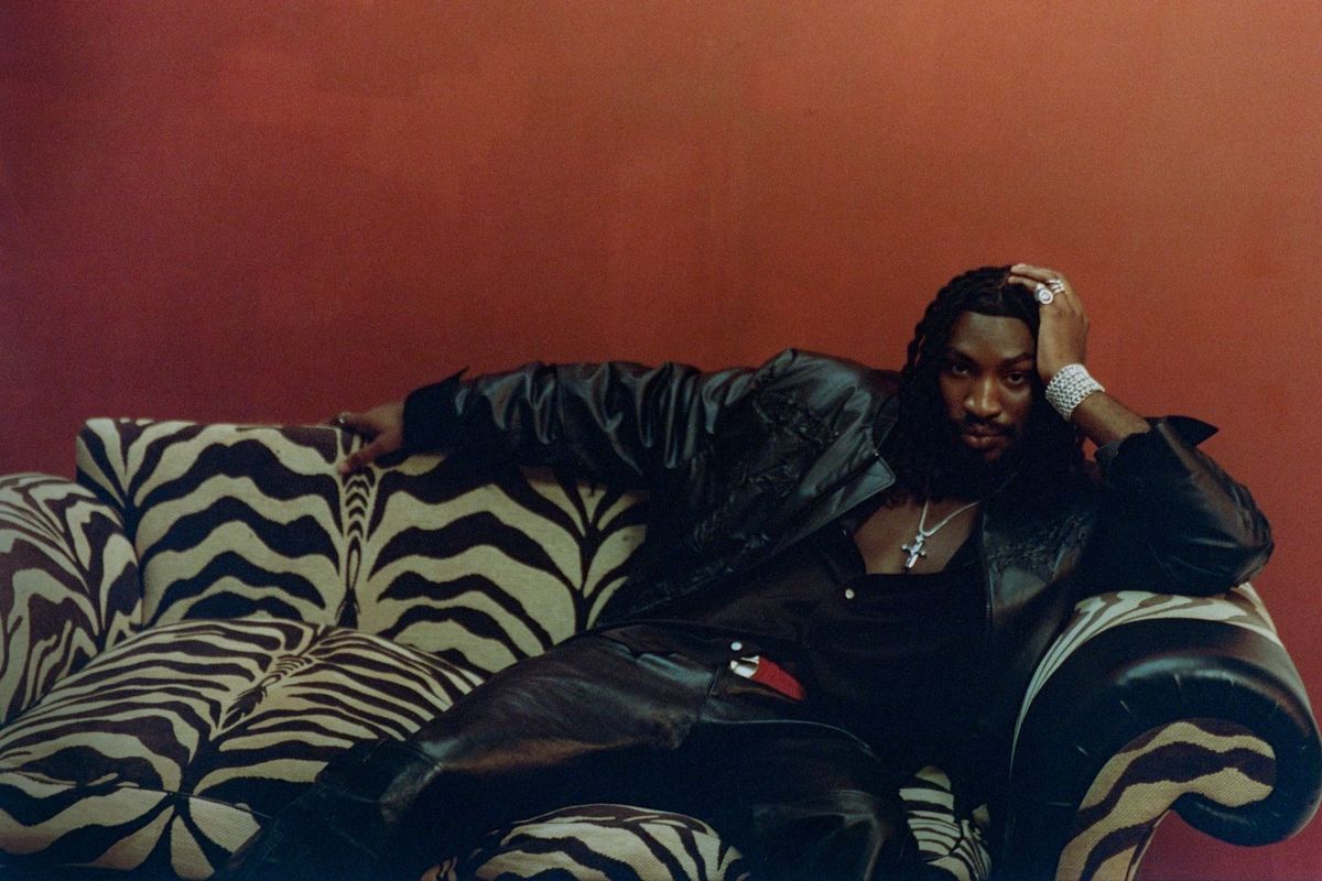 Odeal wears a leather jacket, sitting on a couch of animal hide design
