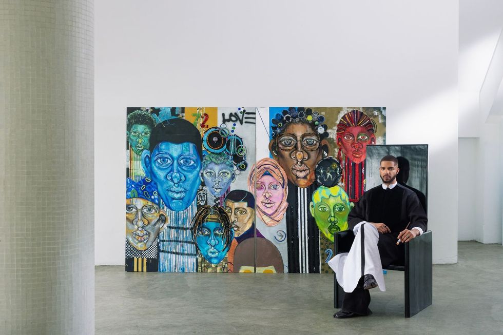 Osoranna sits on a black wood chair, his paintings in the background.