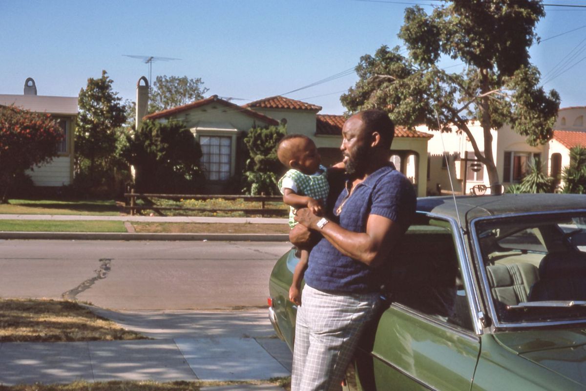 Portrait of an African American man smiling at an African American infant he holds in his arms, he leans against a green car.