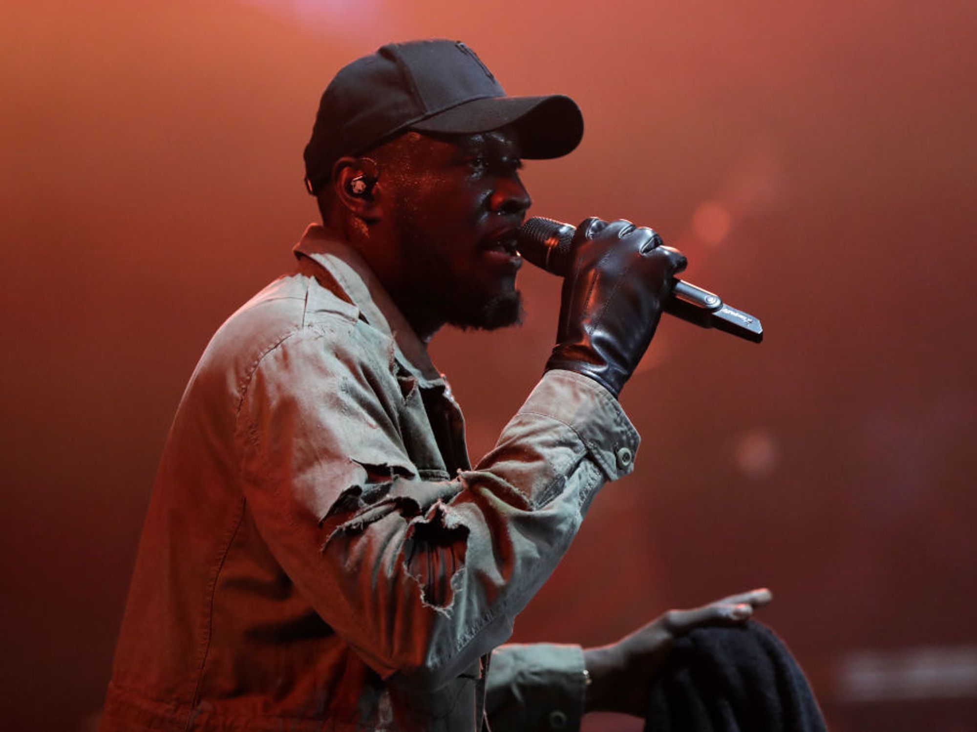 Rapper Stormzy performing with microphone in black hat and beige jacket. 