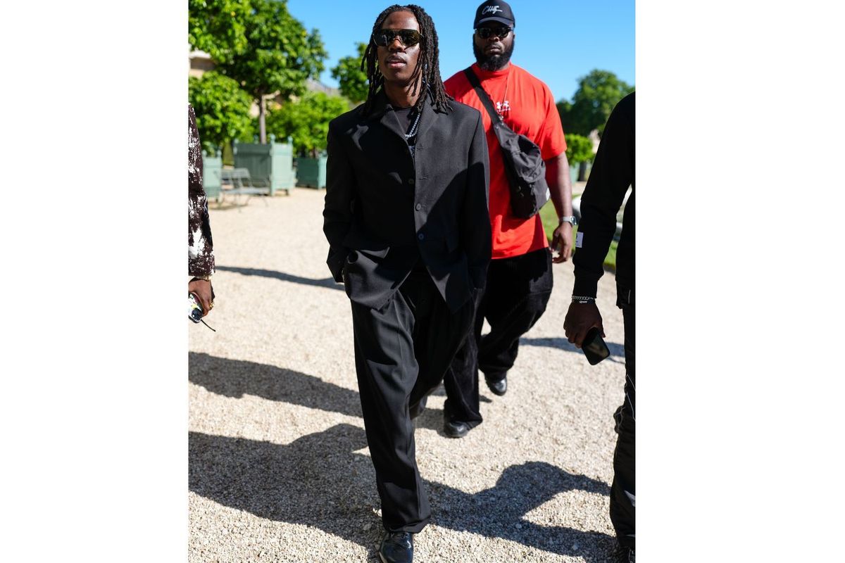 Rema wears a three-piece black tailored suit composed of a structured jacket, buttoned waistcoat, and loose-fitting trousers, paired with polished black leather shoes