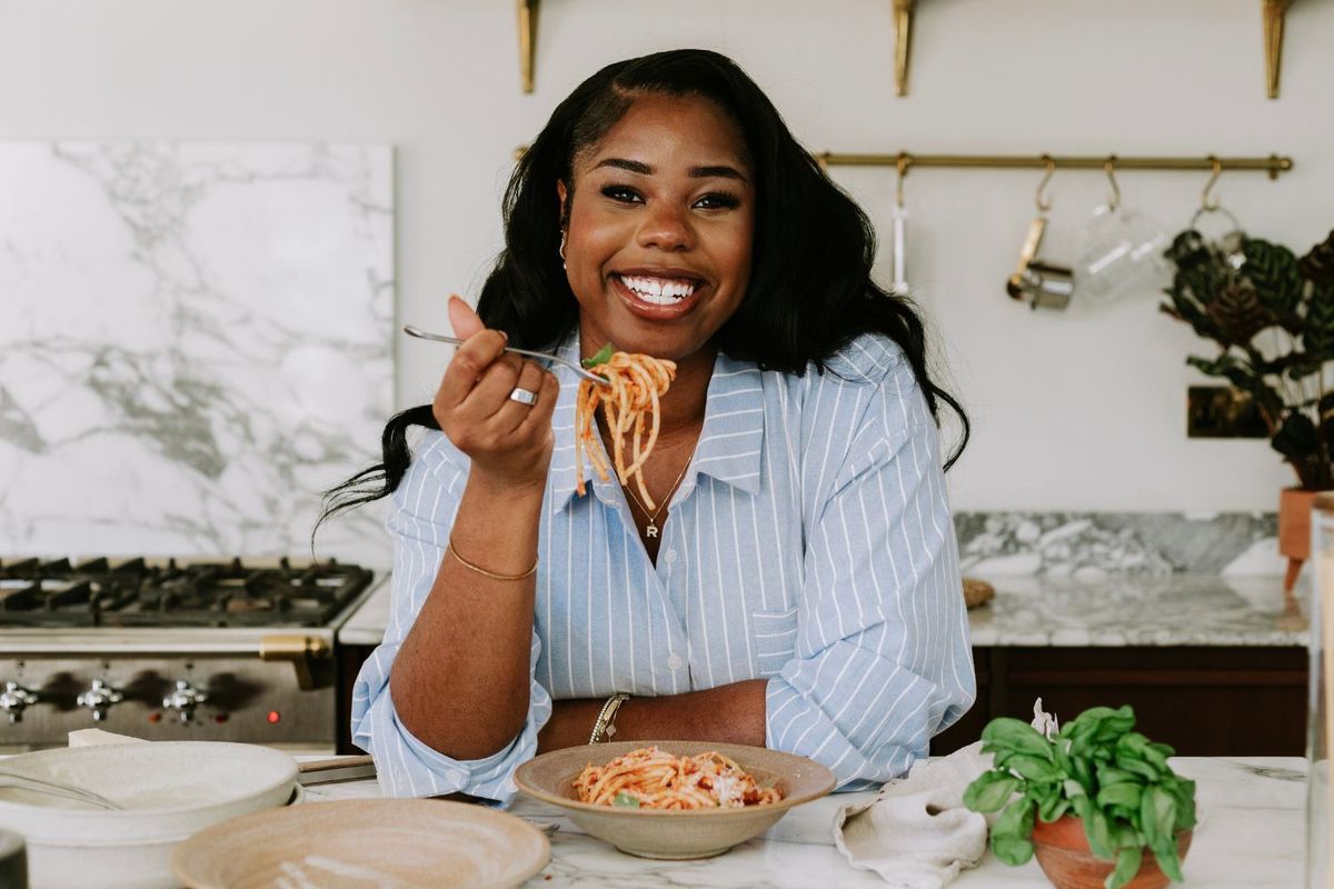 Remi Idowu smiling while eating pasta in a modern kitchen, wearing a blue striped shirt with fresh herbs and cooking ingredients visible on marble countertop.