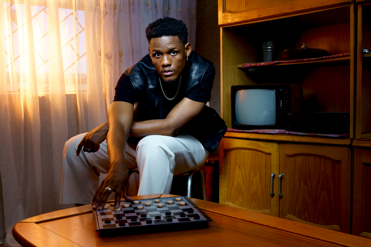 Samthing Soweto makes a move on a board game while gazing at the camera. He wears a black sleeveless leather jacket, black shirt and white pants.