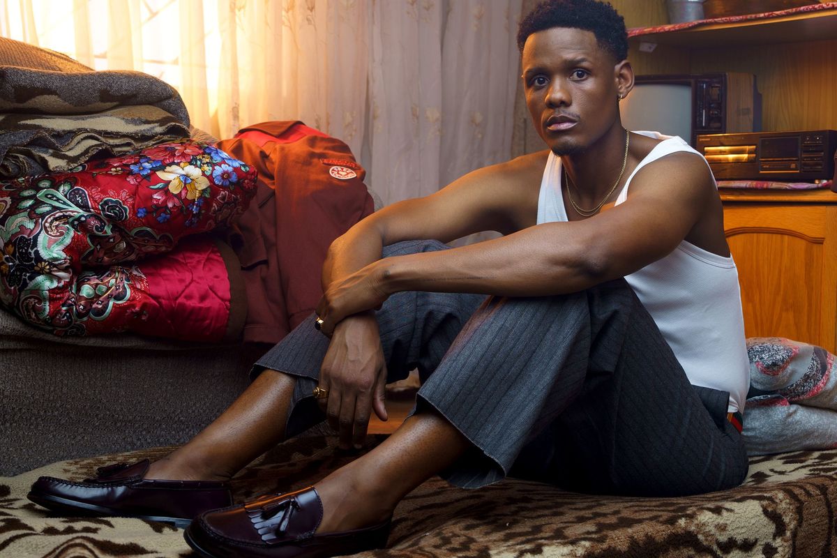Samthing Soweto poses for the camera while seated on a blanket. He wears maroon Dakota shoes, grey pants and a white vest.