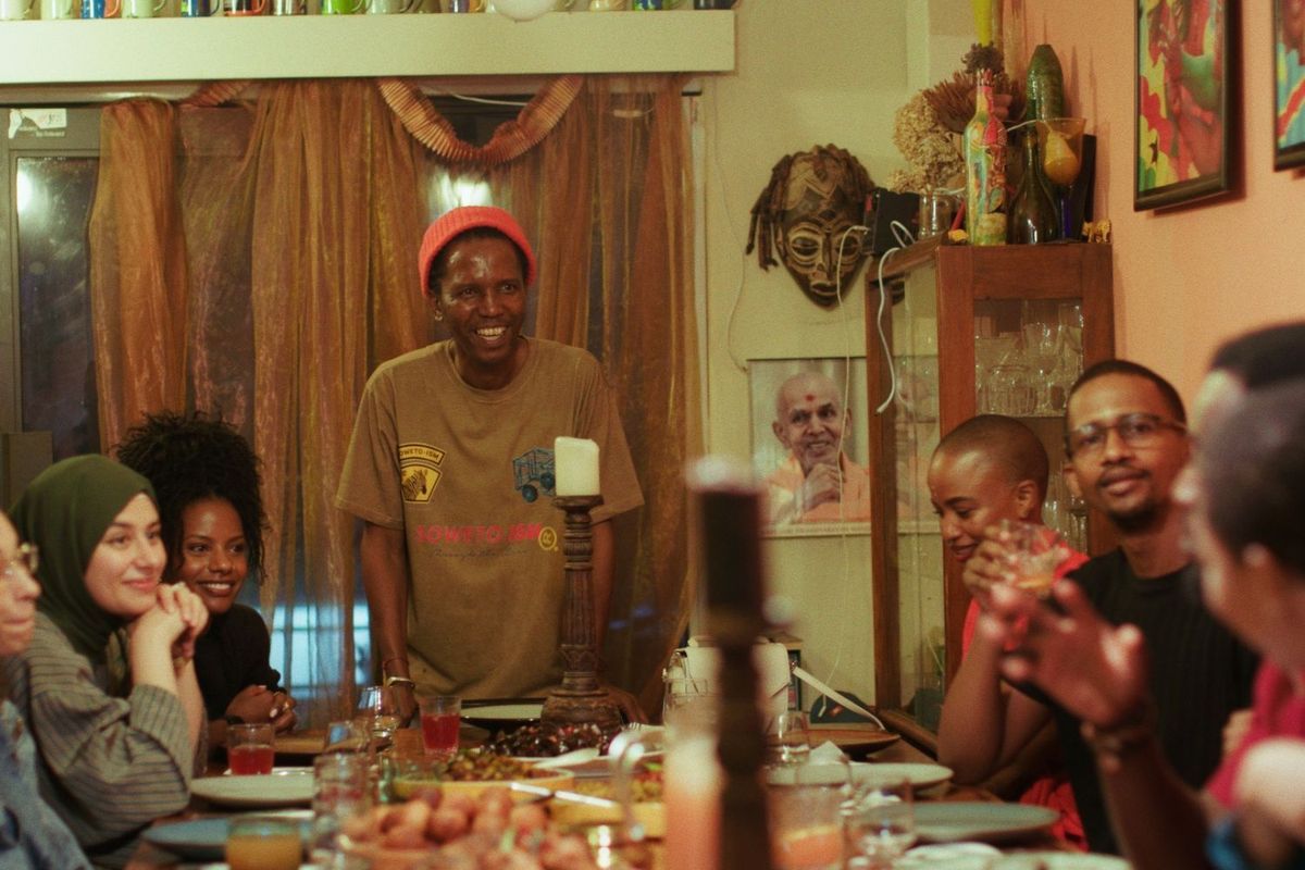 Sanza Sandile hosts guests at his Yeoville Dinner Club. He wears a red woolen hat and a Soweto-ism t-shirt.