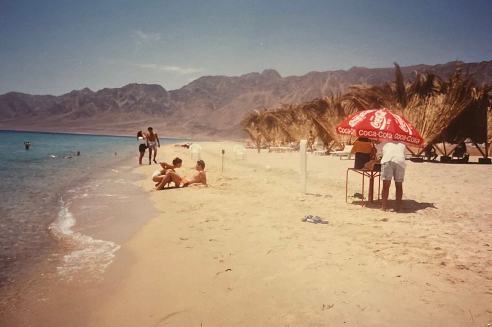 Several people are enjoying themselves on the beach. There's a red Coca-Cola umbrella and some straw huts; the Red Sea Mountains are in the background.