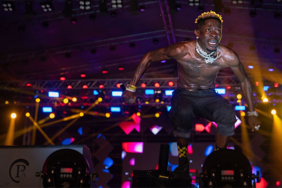 Shatta Wale jumps on stage during a performance; he is shirtless and wearing thick silver chains.