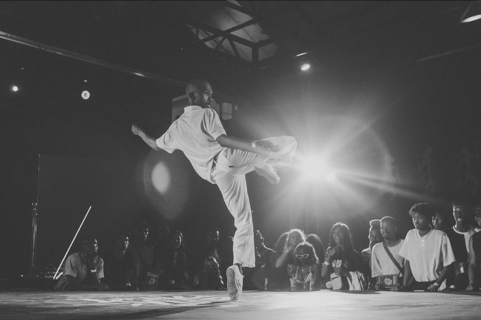 Tebza dances during a dance competition. He is dressed in all white and has one leg raised.