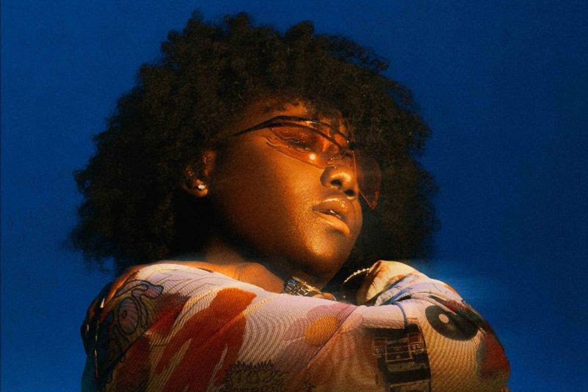 Teni hugs her shoulders in a colorful top against a blue background. 