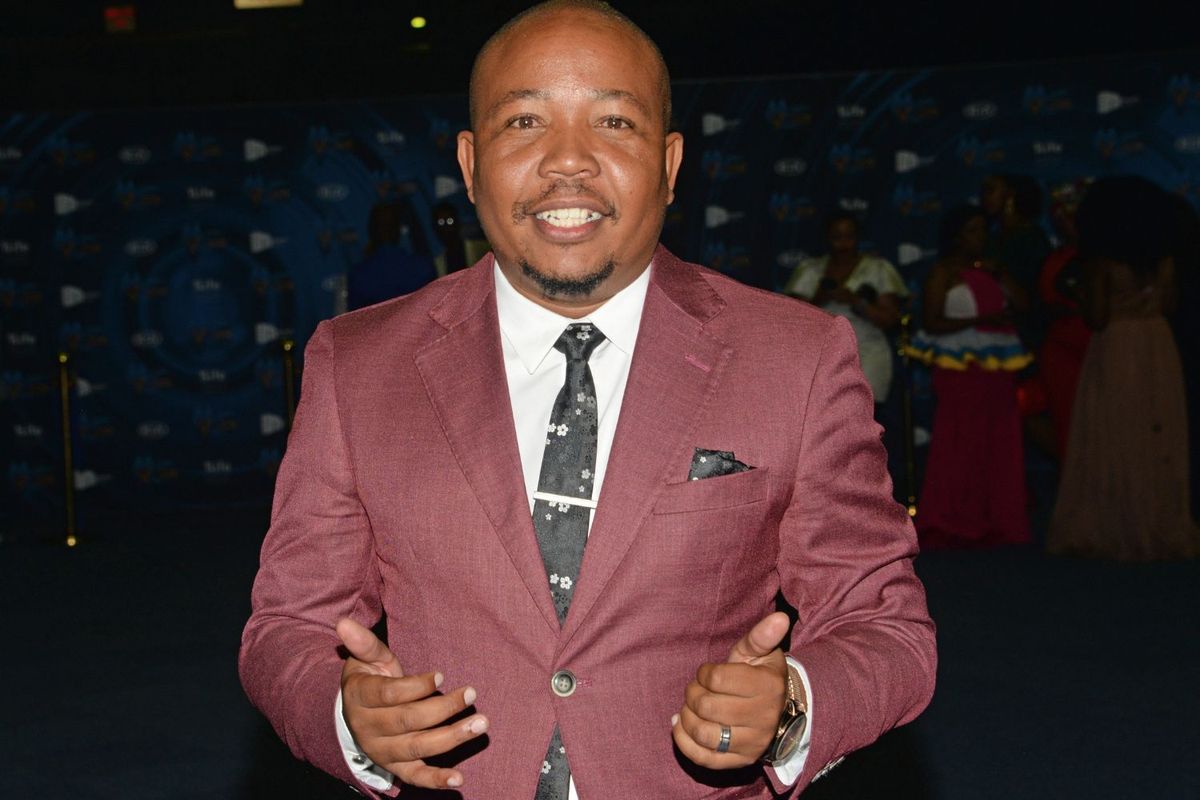 The late South African actor Presley Chweneyagae smiles at the camera. He is in a maroon suit and patterned tie poses for a photo at an indoor event, with people and a branded backdrop visible in the background.