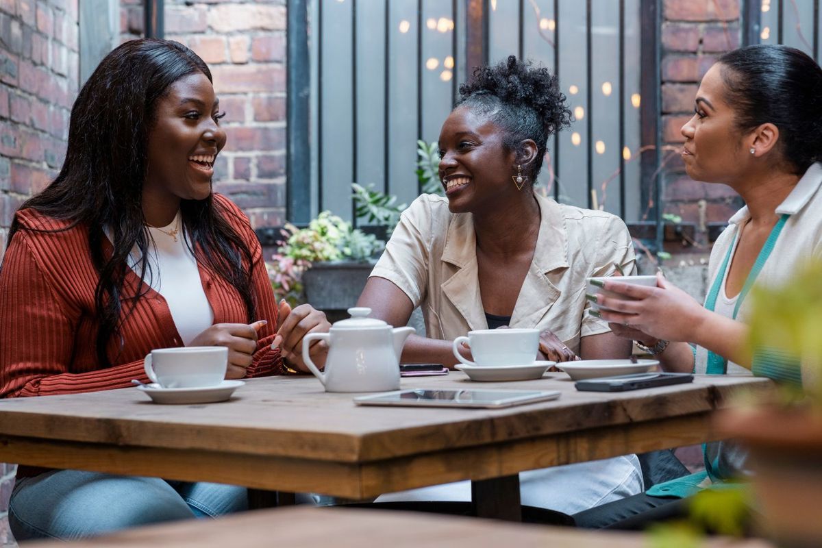 Three women, all smiling, sit together at a wooden café table outdoors, enjoying tea and lively conversation. One woman wears a red cardigan and jeans, another wears a cream short-sleeved jacket with her natural hair in a puff, and the third wears a white blazer with turquoise trim.