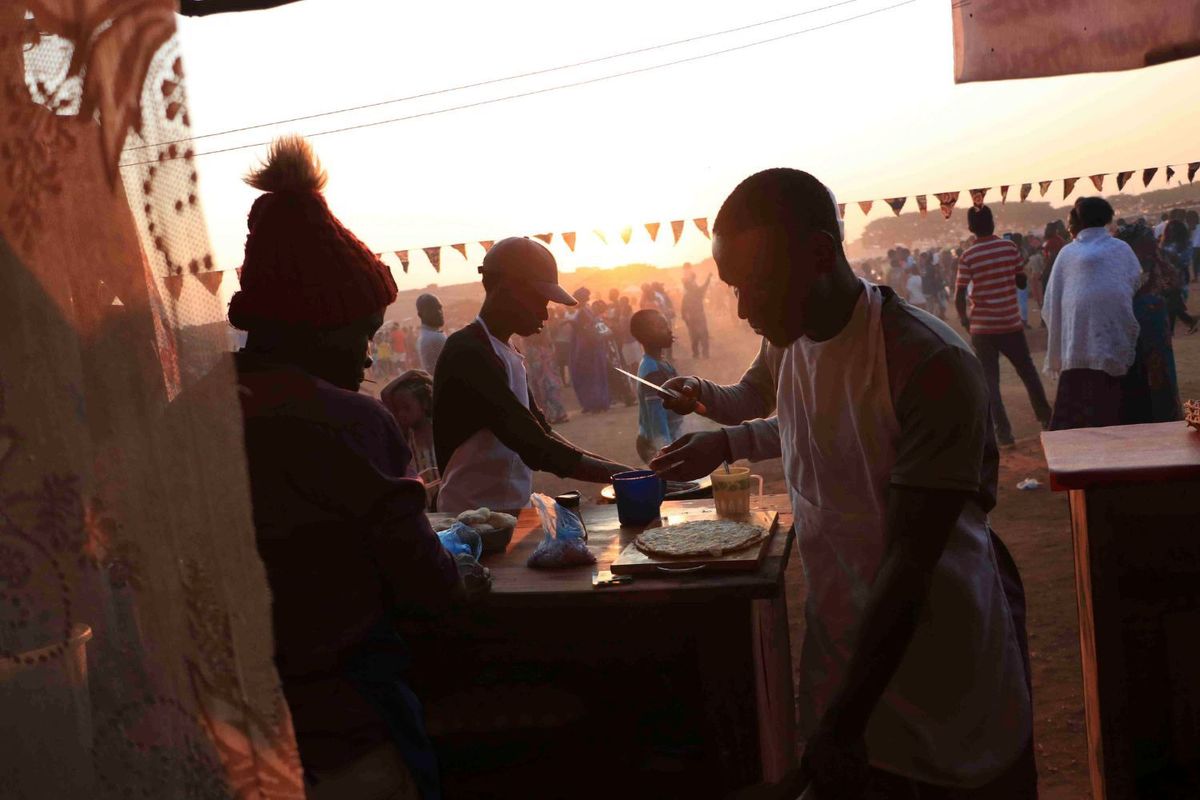 Three young men prepare food on a table with a crowd of people scattered in the background.