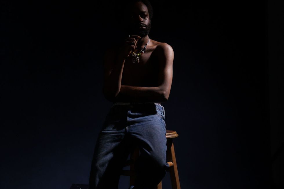 Tim Lyre sits on a bar stool, shirtless and wearing jeans, with a very dark background.