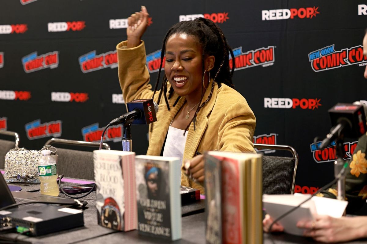 Tomi Adeyemi speaks energetically at New York Comic Con, seated behind a table with fantasy novels and microphones, she is smiling, and her hair is in locks.