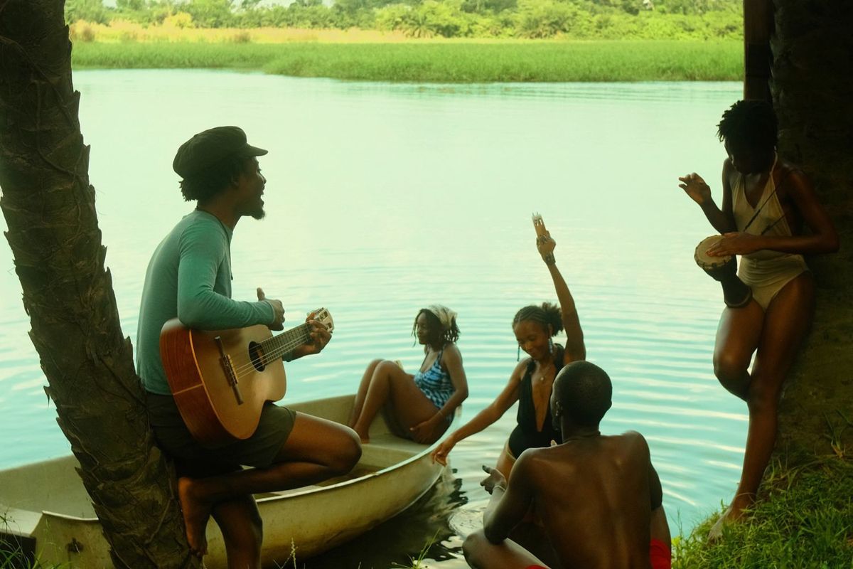 Tommy WÁ is photographed among friends. He is holding the guitar and looking out at the lake, while one woman sits in a boat.
