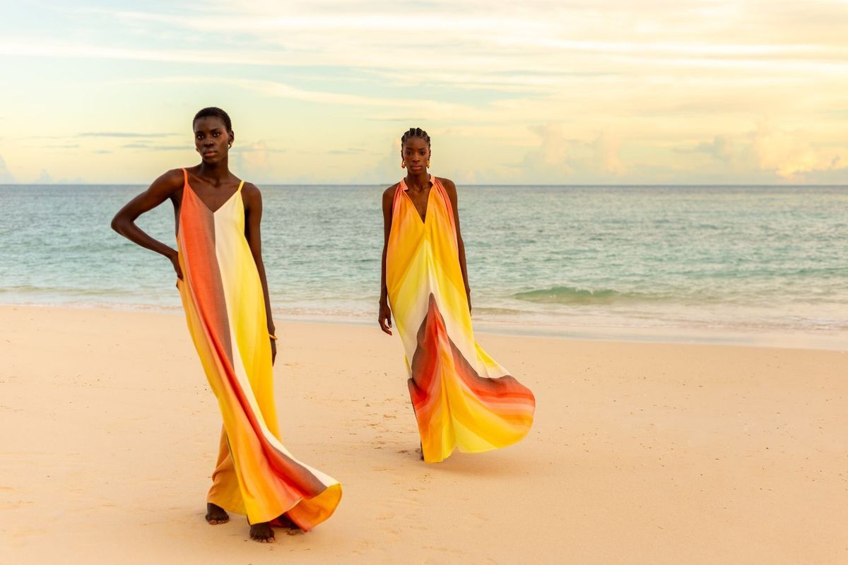 Two models walk barefoot on a beach wearing vibrant yellow, orange, and red striped dresses from Tongoro’s SoLA resort collection, with the ocean in the background.