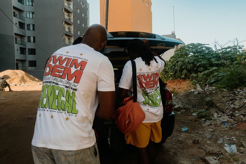 Two people unload the back of a van in white shirts that read "Swim Dem Crew: Senegal."