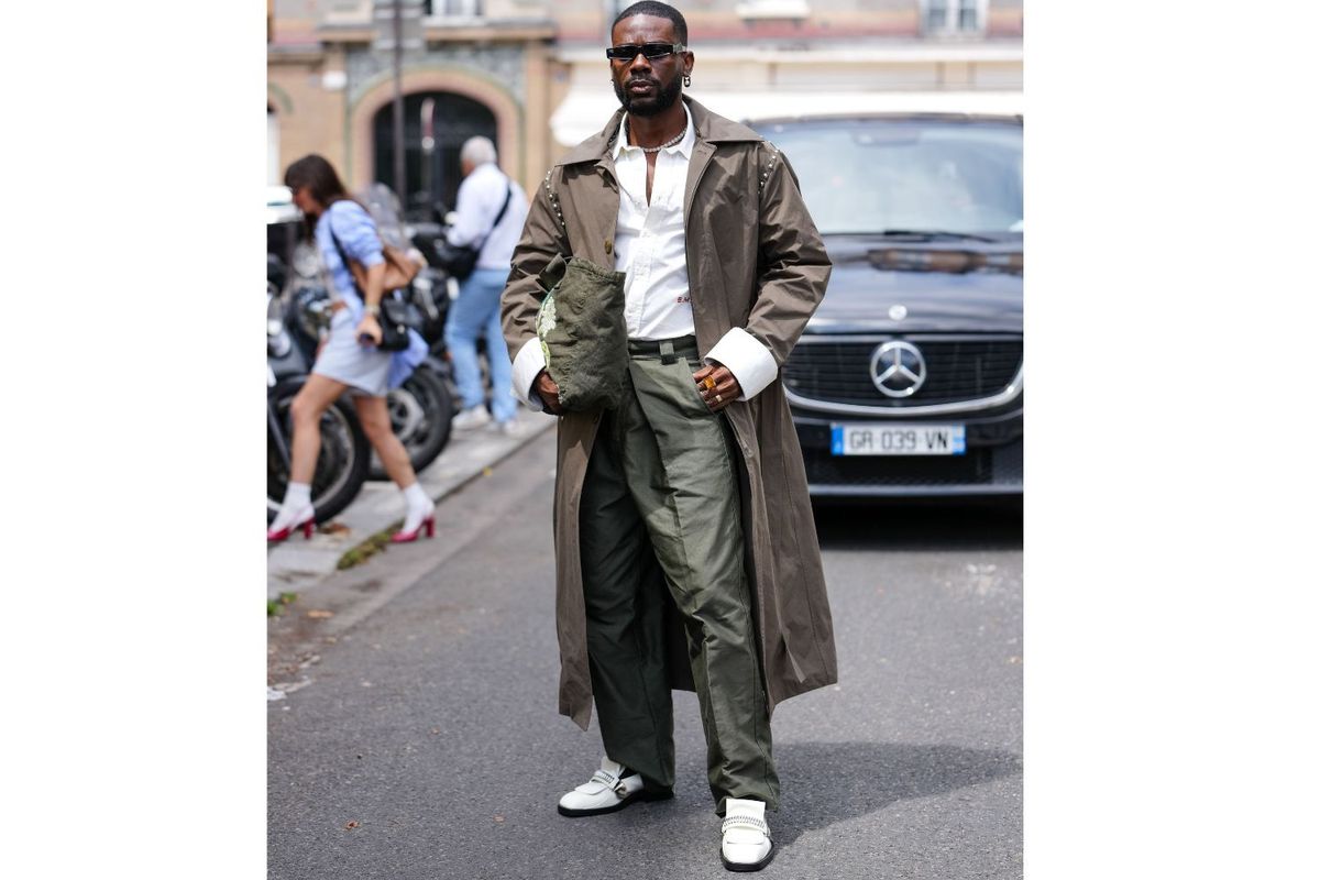 \u200bAdekunle Gold wears silver earrings, white gold diamond choker necklace, white buttoned up long sleeve shirt, dark brown trench coat, dark green tote bag, dark olive green baggy pants, black sunglasses in hand, light yellow gemstone ring, white black Bluemarble loafers shoes