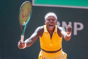Victoria Mboko, wearing a yellow outfit and white headband, celebrates with a yell and clenched fist, holding her racket in the other hand. 