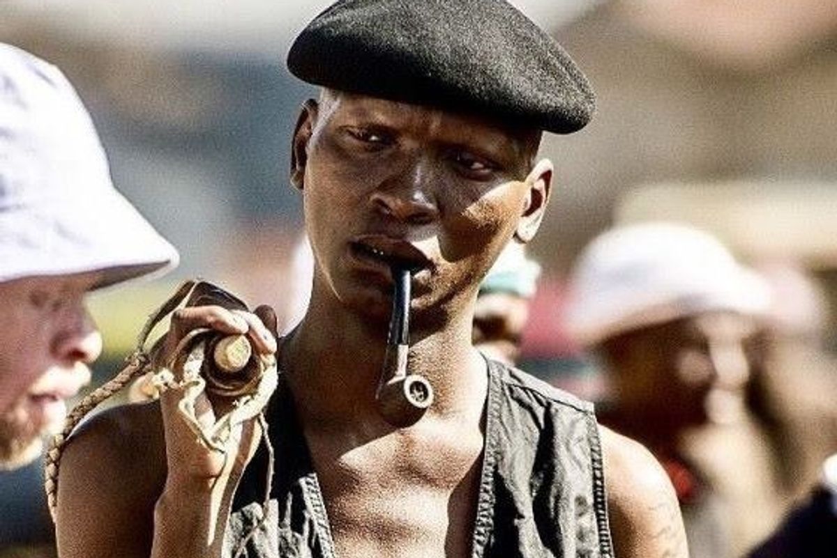 Warren Masemola wearing a black beret with a pipe in his mouth stares intensely at another character from 'Tjovitjo.'